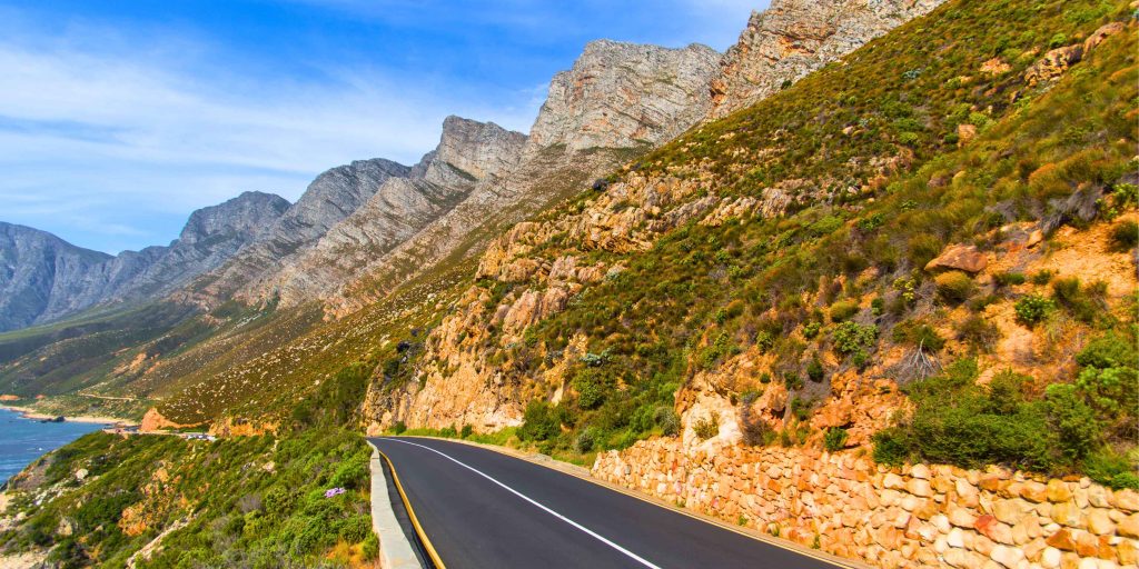 Scenic coastal road winding through rugged mountains overlooking the ocean under a clear blue sky.